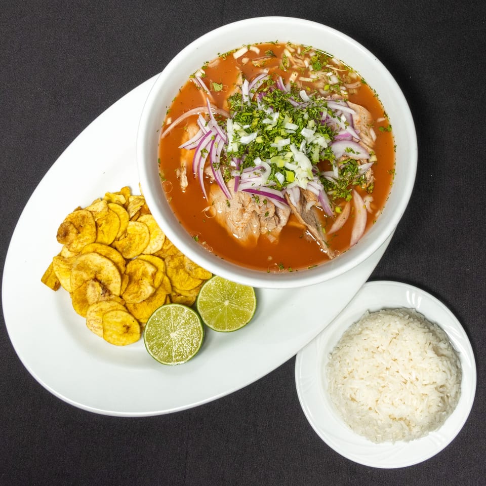 Ecuadorian fish stew with red onions, cilantro, plantain chips, lime, and rice on a black background.